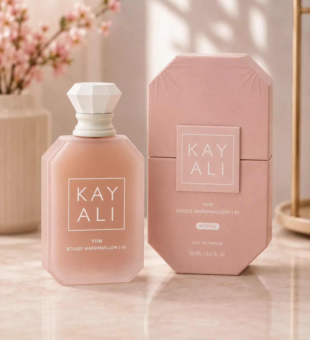 Kay Ali Yum Boujee Marshmallow 81 Eau de Parfum bottle and matching box in a soft blush pink color, displayed on a marble surface with a blurred floral background.