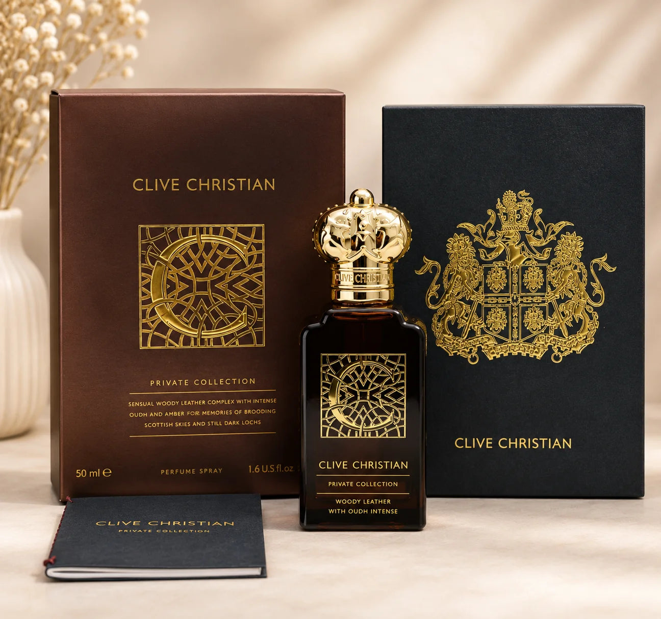 A bottle of Clive Christian Private Collection Woody Leather perfume spray stands in front of its brown box and a black presentation box with gold crest, on a light beige surface with a soft neutral background.