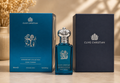 A bottle of Clive Christian Anniversary Collection Iconic Feminine perfume spray stands between two branded boxes on a tabletop. The bottle and box are deep teal-blue with silver '20' and crown graphics, while the right box is dark navy.