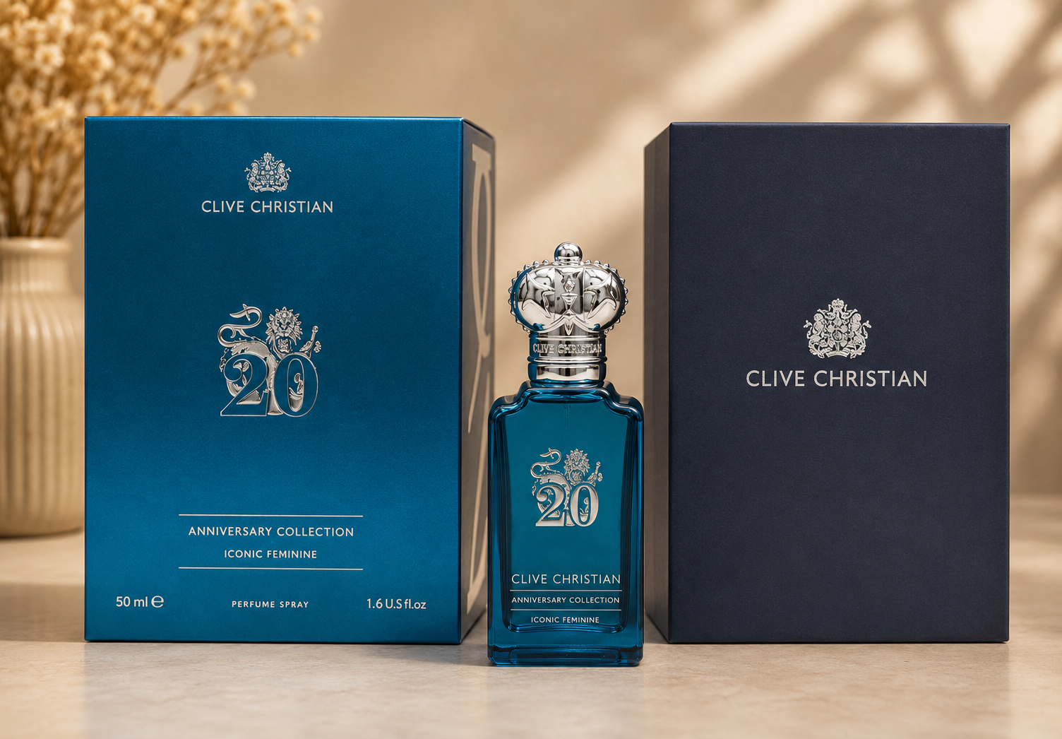 A bottle of Clive Christian Anniversary Collection Iconic Feminine perfume spray stands between two branded boxes on a tabletop. The bottle and box are deep teal-blue with silver '20' and crown graphics, while the right box is dark navy.