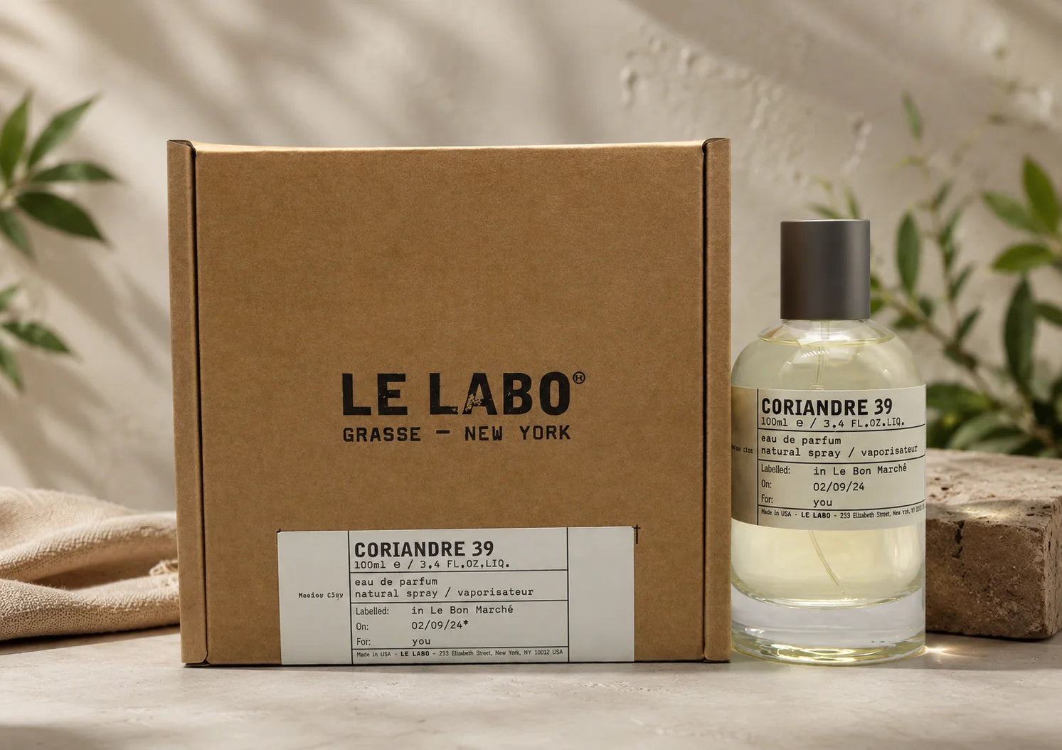 Le Labo Coriandre 39 perfume bottle next to its brown cardboard packaging box, styled on a stone surface with a linen cloth and green leaves in the background.