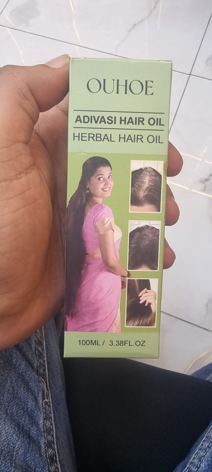 A hand holding a box of OUHOE Adivasi Hair Oil Herbal Hair Oil. The box is light green with text and an illustration of a woman with long dark hair wearing a pink dress.