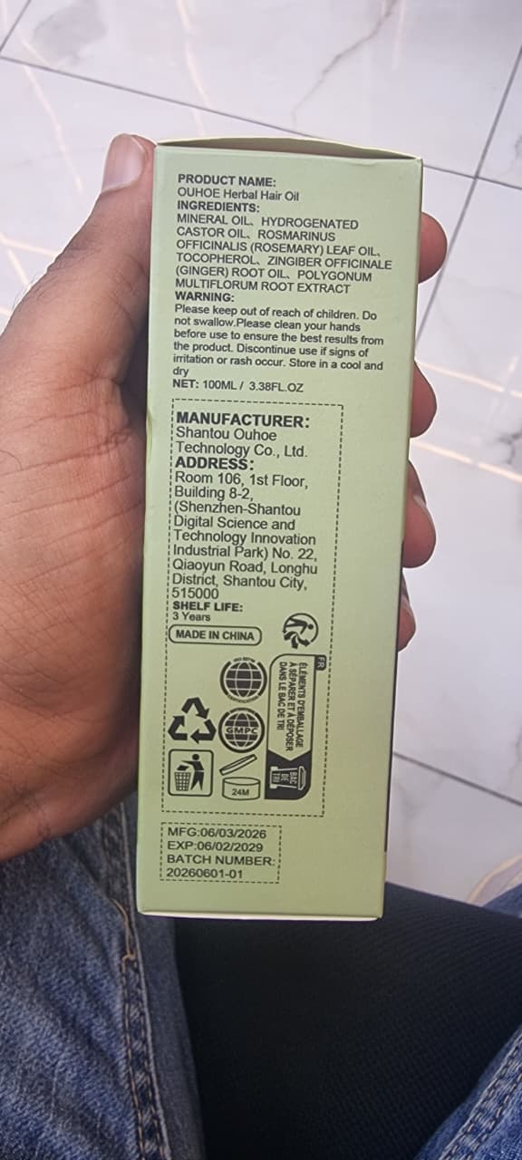 A hand holding a light green rectangular box showing the back side with printed product information, photographed indoors on a tiled floor.